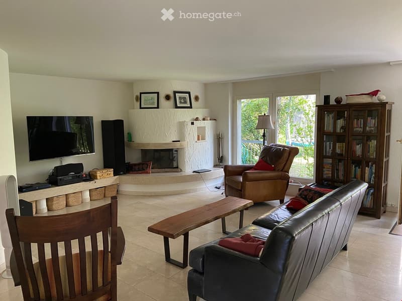 Living room with a fireplace, large windows with a view, a leather sofa, a wooden chair, a wooden table, a bookshelf, and a television.