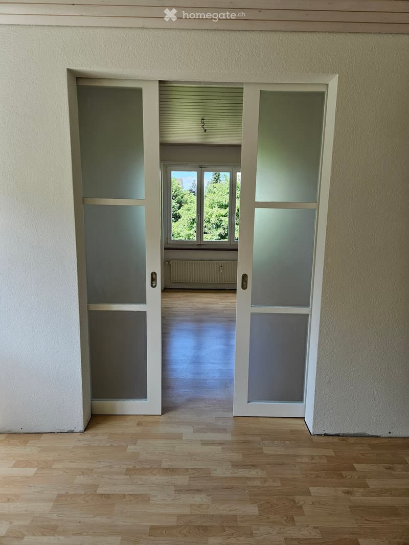 Empty room with wooden flooring, white walls, and large windows with blinds. The room has a door leading to another space with a radiator visible.