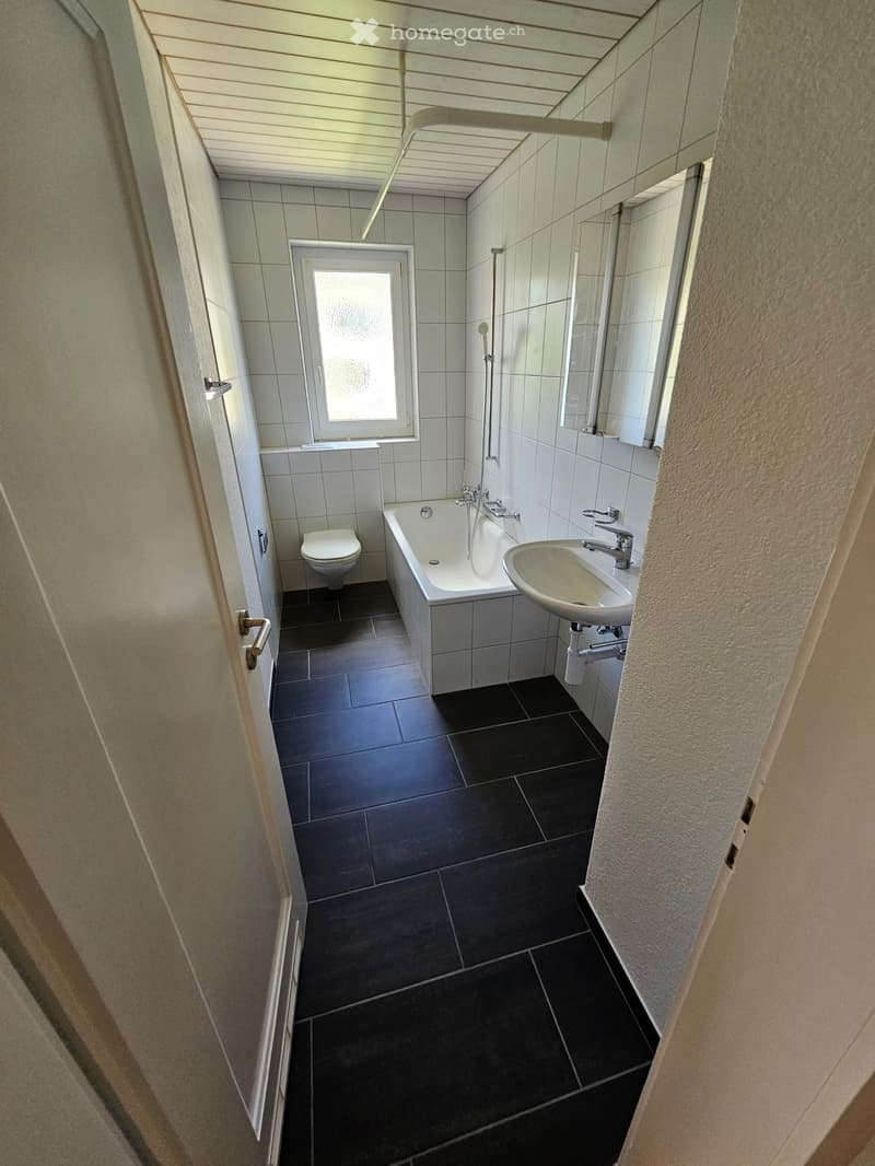 Modern bathroom with black tiled floor, white tiled walls, bathtub, toilet, sink, and window with frosted glass.