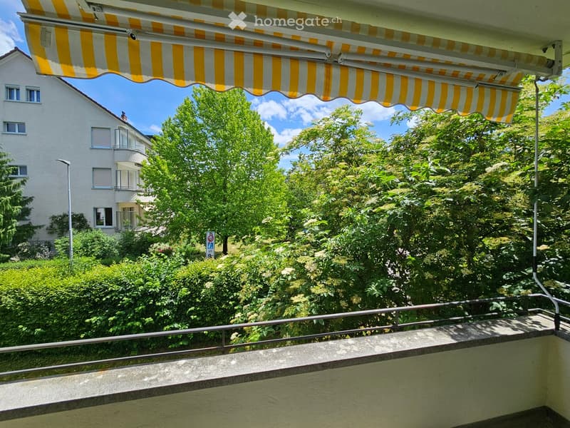 Balcony with a yellow and white striped awning, overlooking a lush green garden and a multi-story residential building.