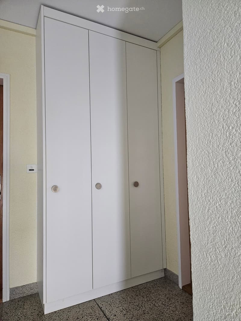 A narrow hallway with white built-in cabinets on one side and textured walls on the other. The floor is tiled, and the ceiling is white.