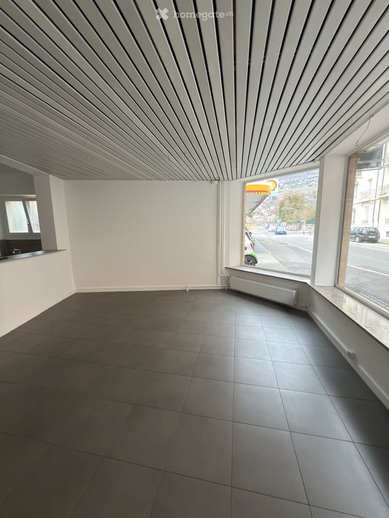 Empty room with grey tiled floor, white walls, large windows, and a radiator. No furniture or equipment present.