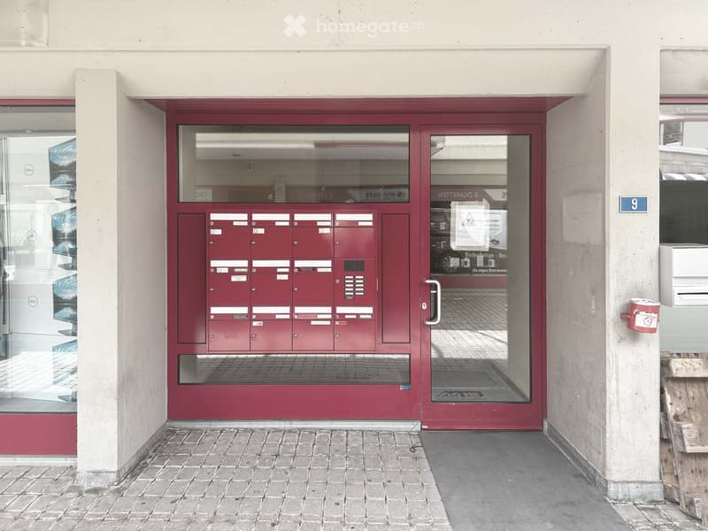 Entrance to a building with a red door, mailboxes, and a glass panel. The number "9" is displayed on the wall to the right.