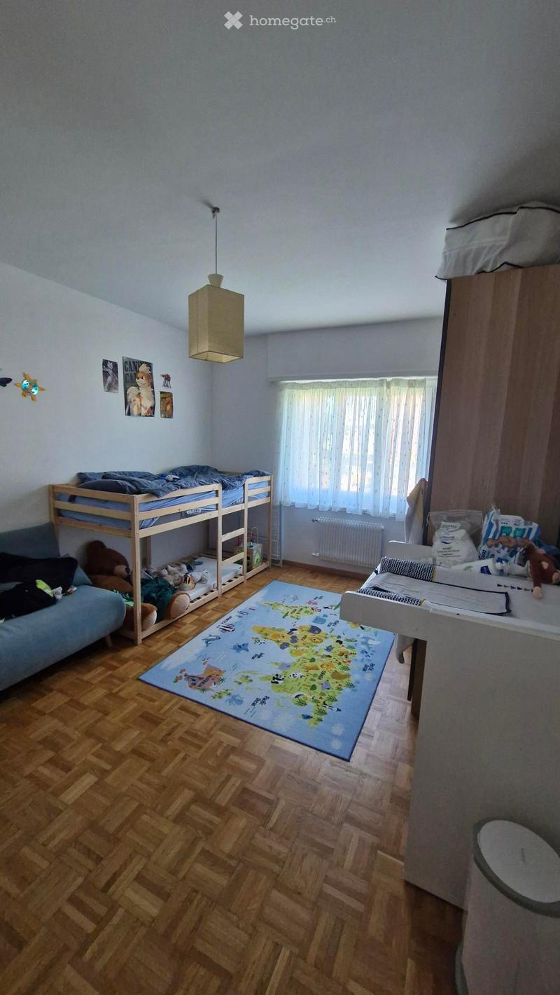 A cozy children's bedroom with a bunk bed, a sofa, a colorful world map rug, and various toys scattered around. The room features wooden flooring and a large window with curtains.
