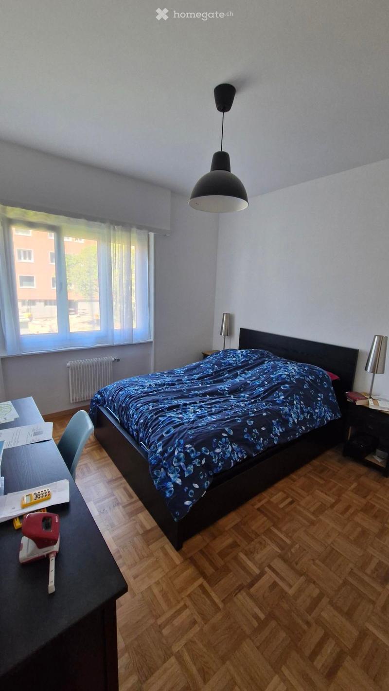 Bedroom with wooden flooring, a bed with a blue floral duvet, a nightstand with a lamp, a desk with a chair, and a window with blinds.