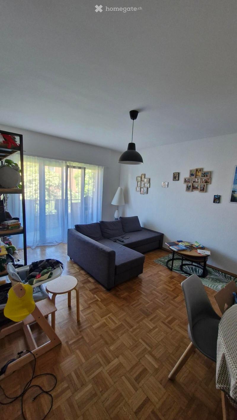 Living room with wooden herringbone flooring, grey sectional sofa, coffee table, small round table, and various seating options including a chair and stool. Shelving unit with books and decorative items, and wall-mounted picture frames.