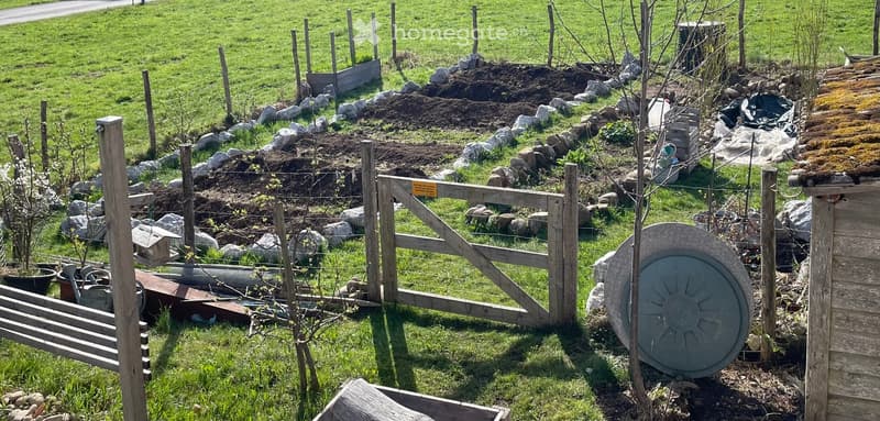 A well-maintained garden area with a wooden gate, raised garden beds, rocks, and a small shed. The area includes a variety of plants and a circular metal object near the gate.