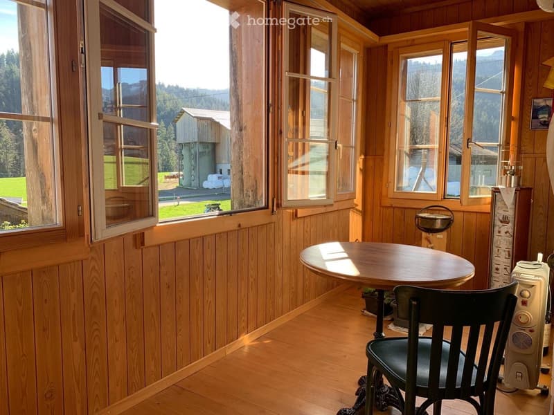 A cozy wooden room with large windows offering a scenic view, featuring a round wooden table, a black chair, and a portable heater.
