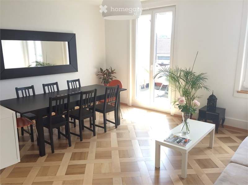 Bright, open living and dining area with wooden flooring, large mirror, dining table with chairs, coffee table with vase, and plants near the window.