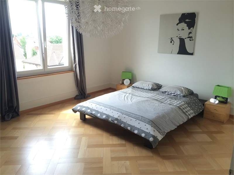 Cozy bedroom with wooden flooring, large window with view, bed with patterned bedding, nightstands with lamps, and a stylish picture frame on the wall.