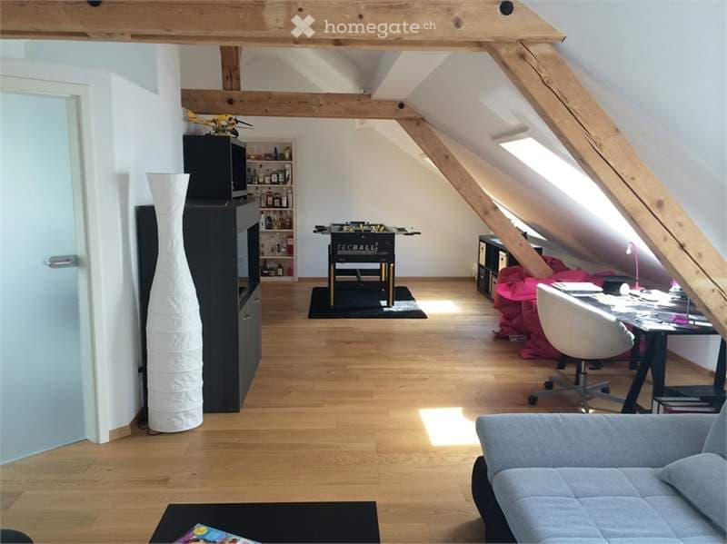 Bright, open attic space with wooden beams and exposed ceiling. Features a grey sofa, coffee table, desk with chair, bookshelf, tall decorative vase, and a foosball table.