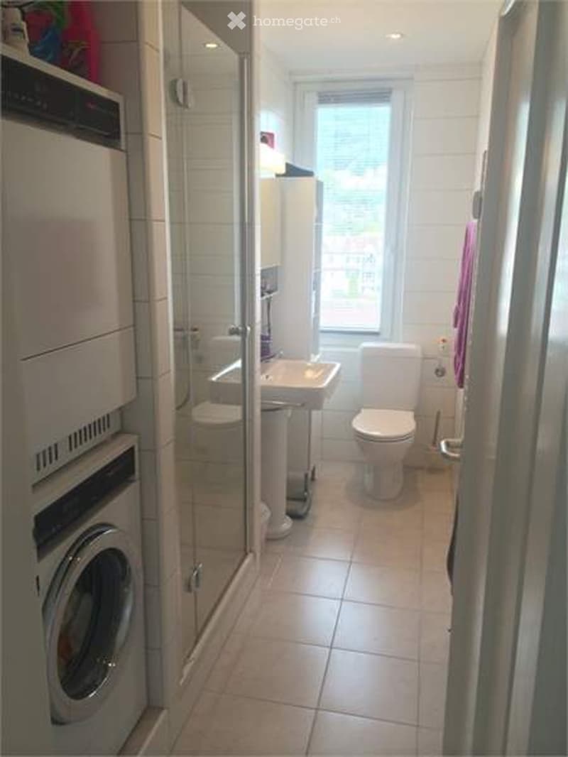 Modern bathroom with white tiles, a toilet, sink, shower, and a washing machine on the left side.