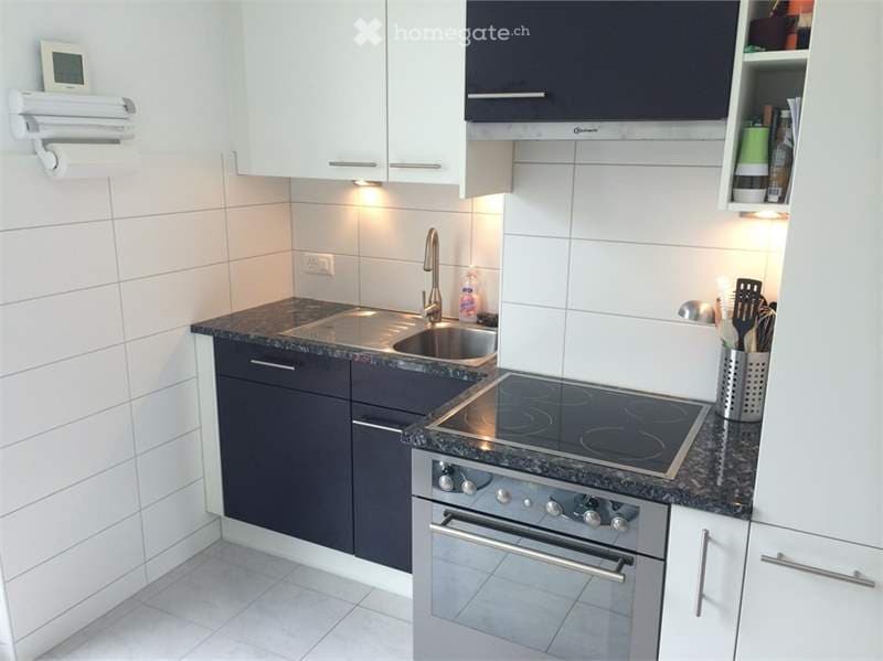 Modern kitchen with white cabinets, dark countertops, stainless steel sink, induction cooktop, and oven. Includes various kitchen utensils and a paper towel holder.