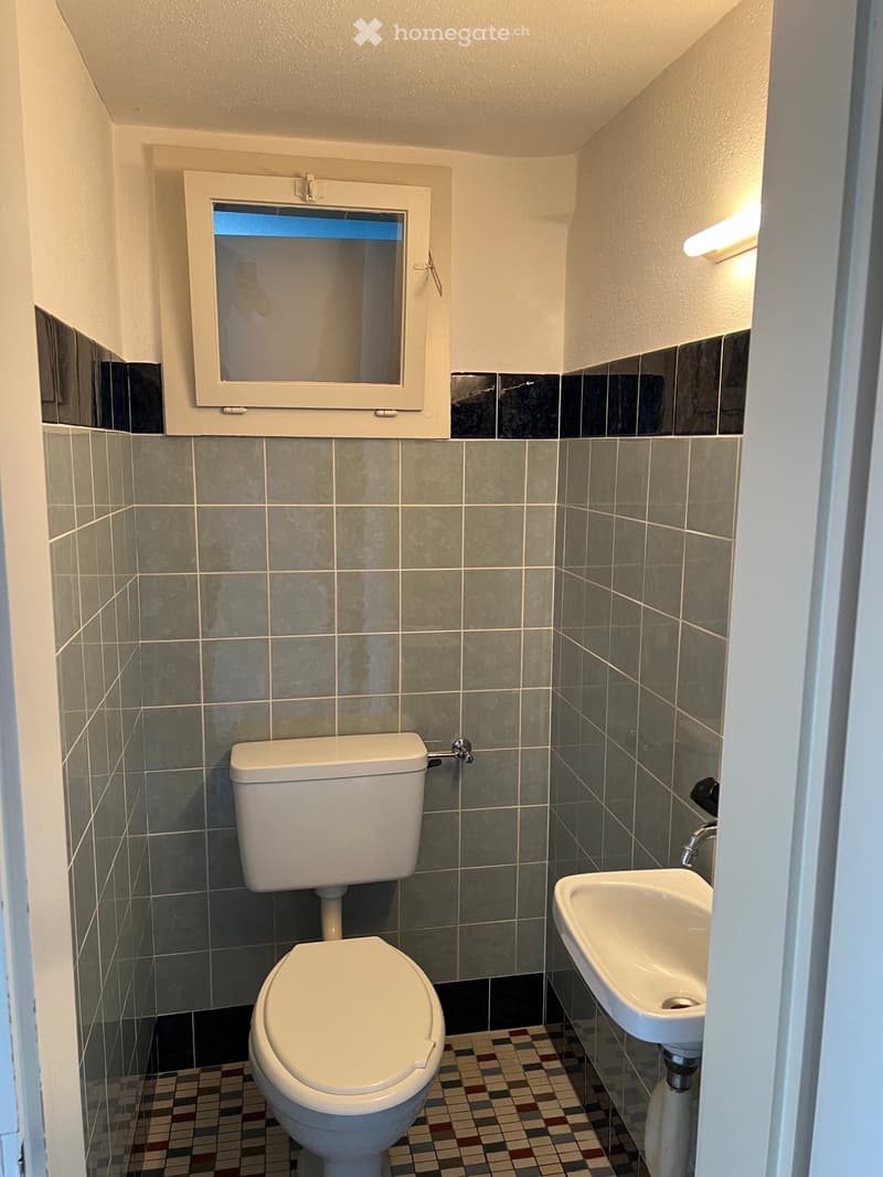 Small bathroom with grey tiled walls, a toilet, a sink, and a window above the toilet.