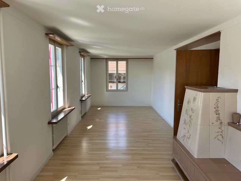 Empty hallway with wooden flooring, white walls, and large windows allowing natural light. Radiators are visible along the walls, and there is a small cabinet with floral designs on the right side.