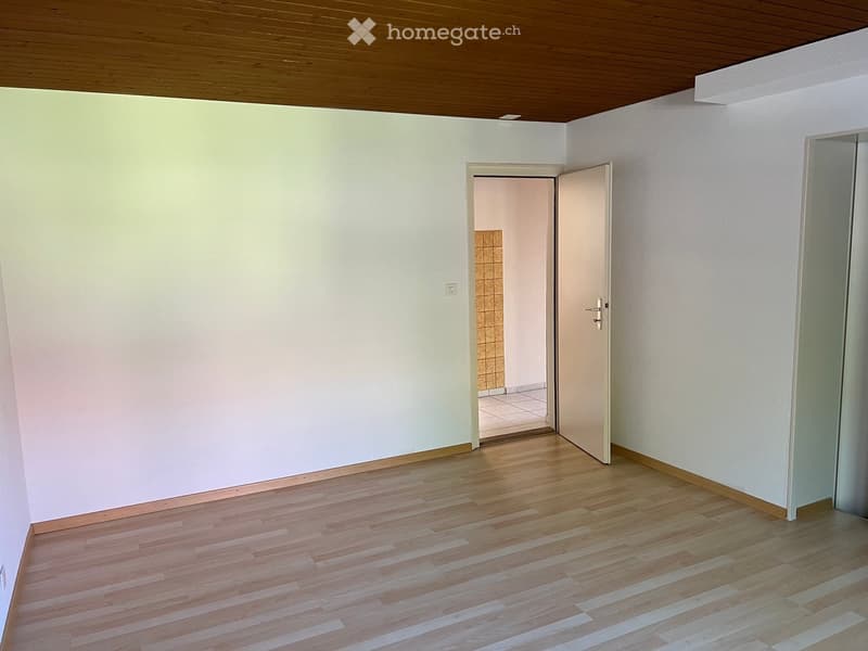 Empty room with light-colored walls, wooden flooring, and a wooden ceiling. An open door leads to another room with tiled walls and flooring.