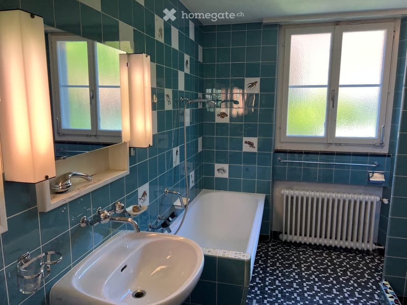 Bathroom with blue tiles, a sink, bathtub, radiator, and a mirror. The room has two windows and a glass shelf with a cup.