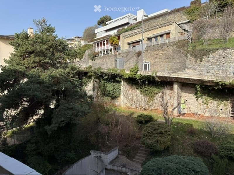 Multi-story building with raised ground floor, stone walls, and greenery in the foreground.
