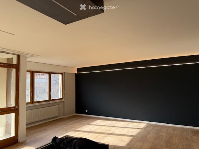 Empty room with wooden flooring, large windows, and dark accent wall. No furniture or equipment present.