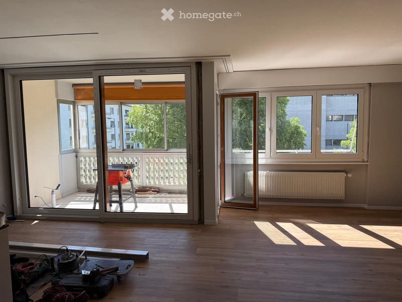 Empty room with wooden flooring, large windows, and a sliding glass door leading to a balcony area. Tools and equipment are visible on the floor near the entrance.
