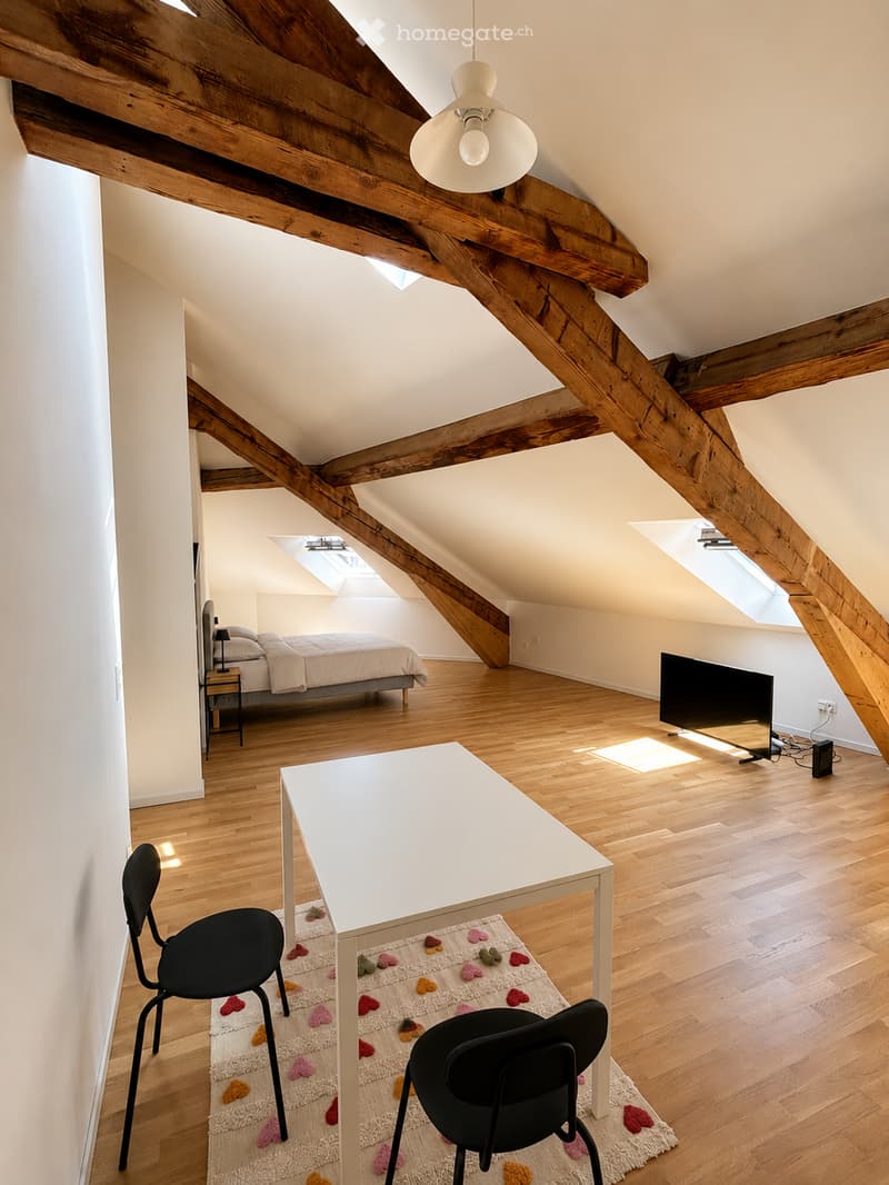Bright, airy attic bedroom with wooden beams, hardwood floors, and large skylights. Features a bed, nightstand, desk with chairs, and a flat-screen TV.