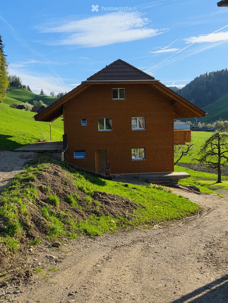 A wooden house with a sloped roof, multiple windows, and a small balcony, situated in a green, hilly landscape.