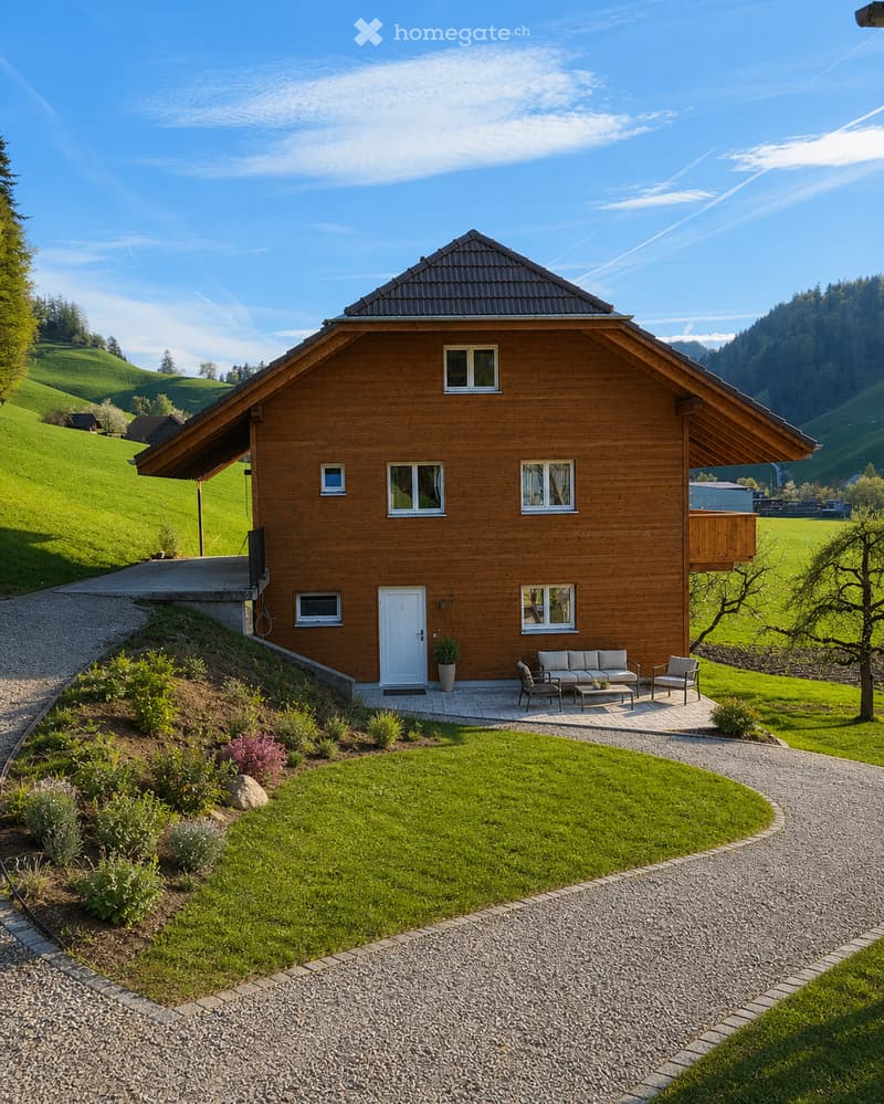 A two-story wooden house with a sloped roof, white door, multiple windows, and a small balcony. The house is surrounded by a well-maintained lawn, garden, and gravel driveway.