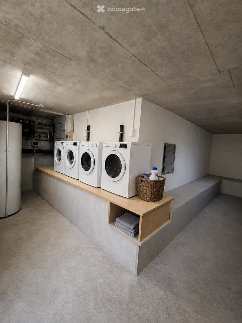 A modern, minimalist laundry room with four white washing machines and dryers arranged on a wooden shelf. A wicker basket with cleaning supplies sits on the shelf, and folded towels are stored in a compartment below.