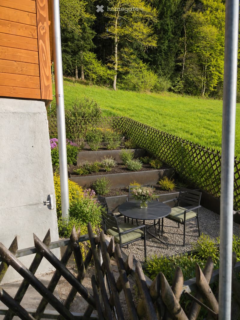 Garden area with a small seating set including a round table and four chairs, surrounded by a wooden fence and lush greenery.
