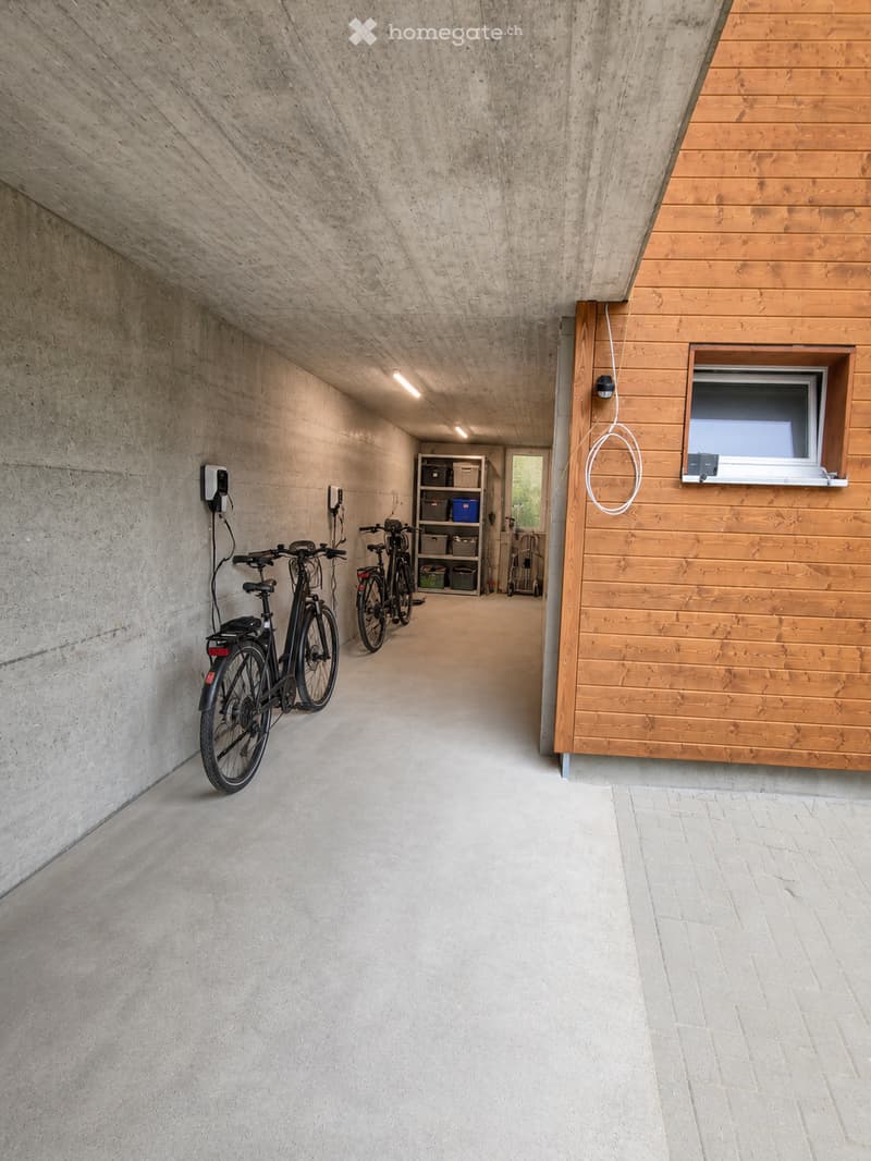 Modern, minimalist corridor with concrete walls and ceiling, wooden paneling on one side, bicycles parked, small shelving unit, and a window with blinds.