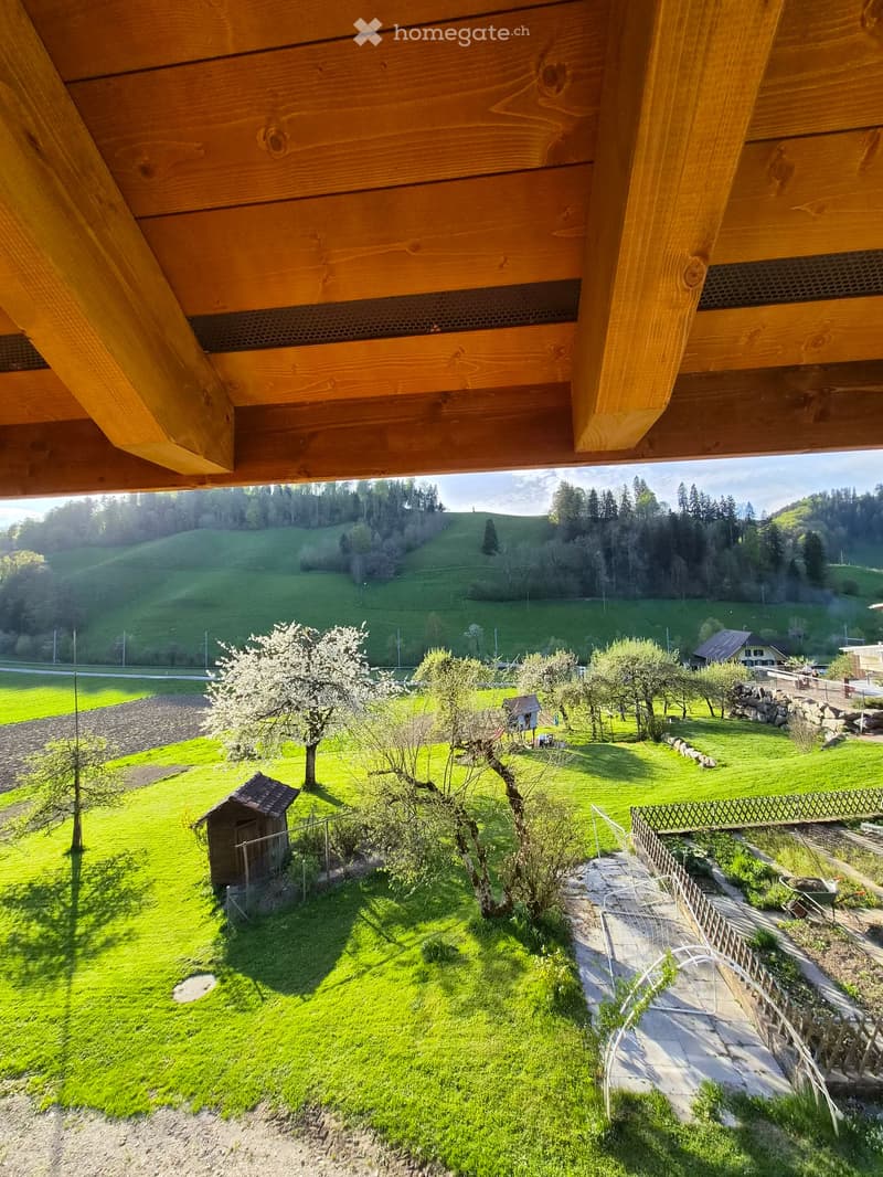 View from a wooden balcony overlooking a lush green garden with trees, a small shed, and a stone fence. The landscape includes rolling hills and a distant house.
