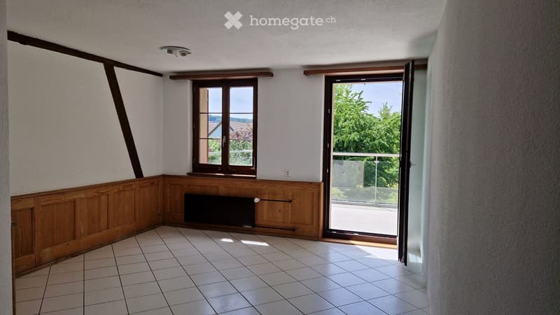Empty room with tiled floor, wooden paneling, and large windows offering a view of greenery. The room features a radiator and a sliding glass door leading to a balcony.