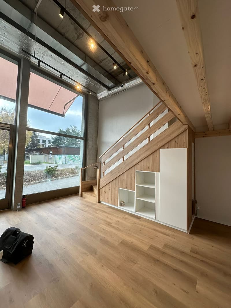 Empty room with wooden flooring, white walls, a staircase with wooden railings, and large windows offering a view of the outside. A small built-in shelf unit is visible on one wall.
