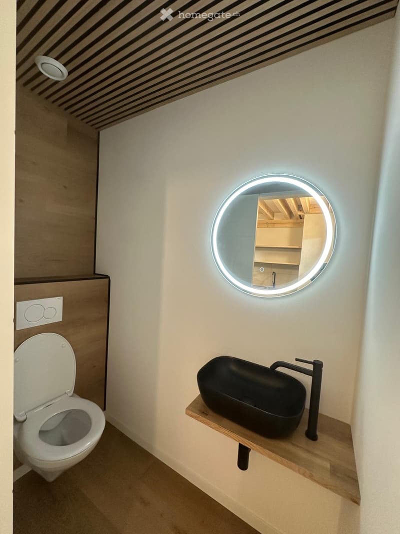 Modern bathroom with a sleek black sink, wooden shelf, round illuminated mirror, and a toilet.