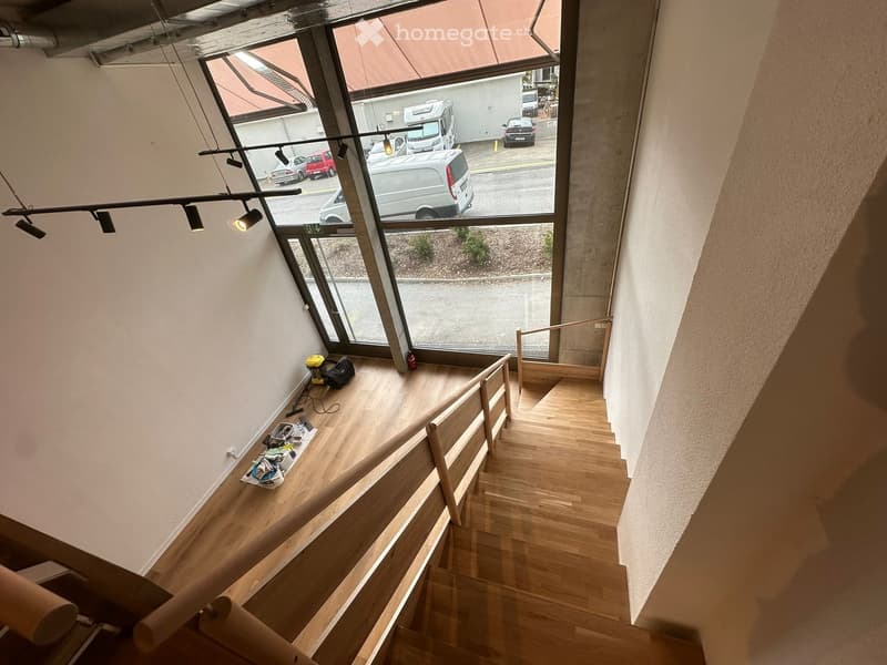 Staircase with wooden steps and railings, large windows overlooking a parking area with several cars, and some tools and equipment placed on the landing.