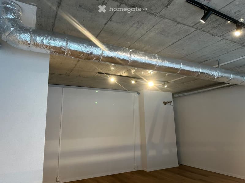 Empty room with wooden flooring, white walls, and exposed wooden ceiling with lighting fixtures. There are two doors and visible air ducts running across the ceiling.