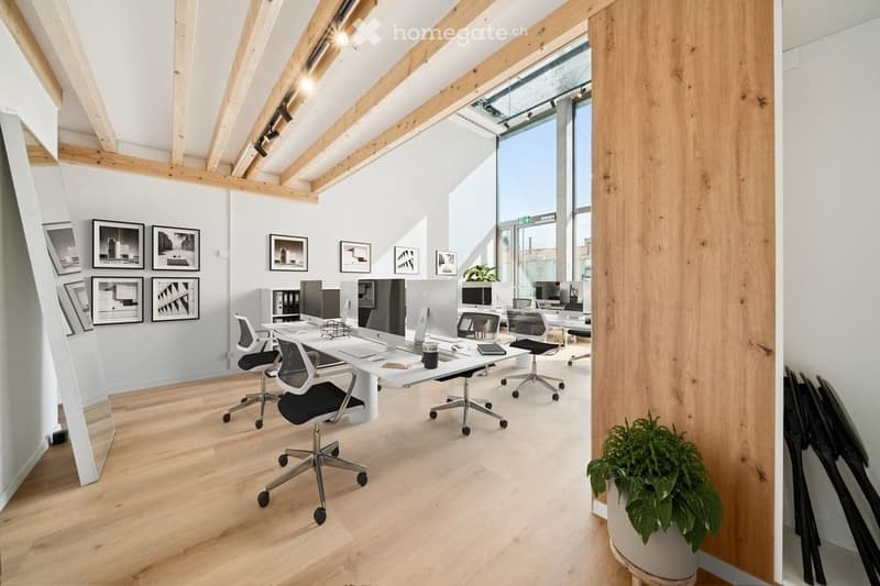 Modern office space with light wood flooring, white walls, exposed wooden beams, large windows, desks, chairs, computers, and framed artwork.