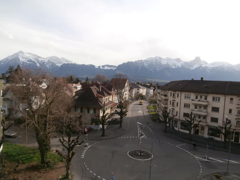 A town square with buildings, trees, and a view of snow-capped mountains in the background.