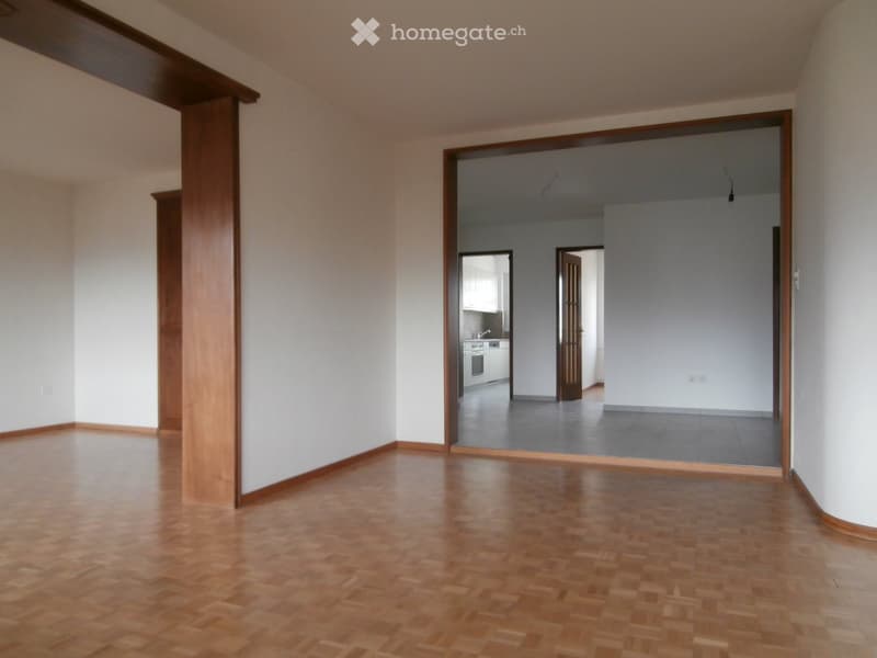 Empty room with wooden flooring and white walls, featuring an open doorway leading to another room with a kitchen area visible in the background.