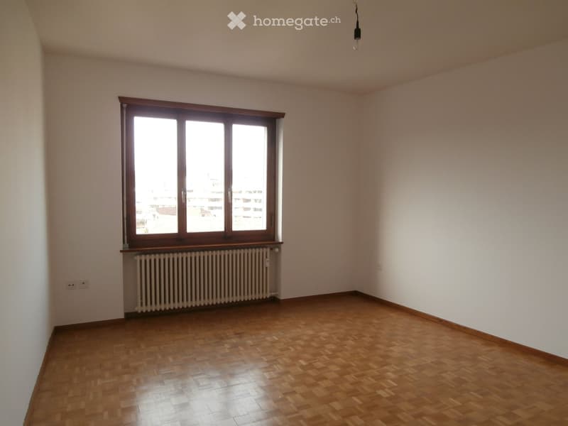Empty room with wooden flooring, white walls, large window with wooden frame, radiator, and visible electrical outlet.