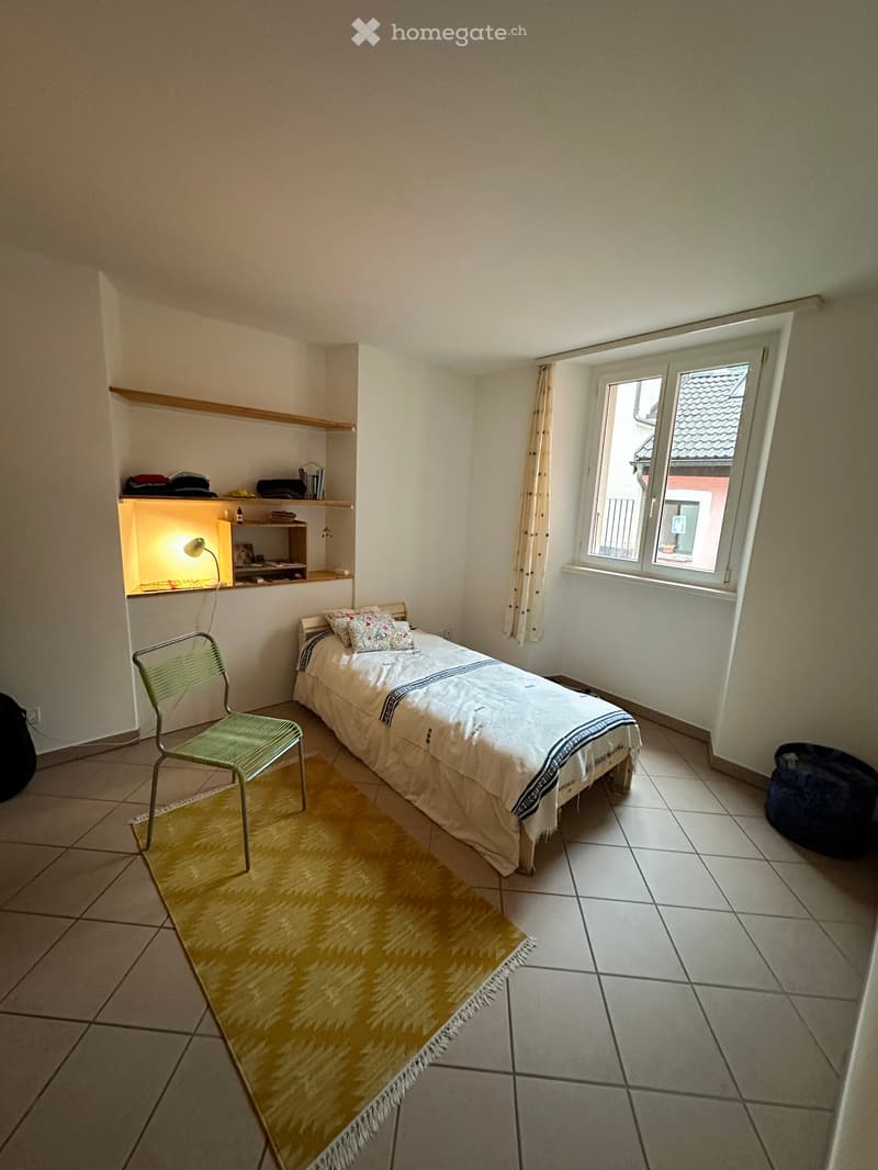 A small bedroom with a single bed, a green chair, a built-in shelf unit with a lamp, and a patterned rug on the tiled floor.