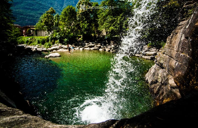 A natural outdoor setting featuring a small waterfall cascading into a clear, green pool surrounded by rocks and lush greenery. A few people are visible near the water, enjoying the scenic environment.