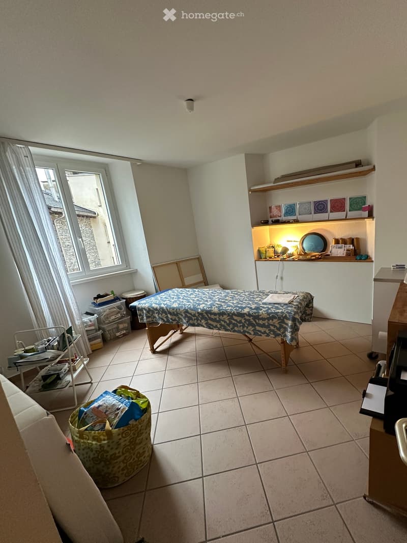 A furnished room with a massage table, shelves, a sofa, and storage bins. The room has tiled flooring and large windows with curtains.