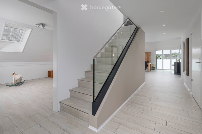 Modern hallway with light wooden flooring, white walls, and a glass-enclosed staircase leading upwards. Features include recessed ceiling lights, a skylight window, and a rocking chair in the corner.
