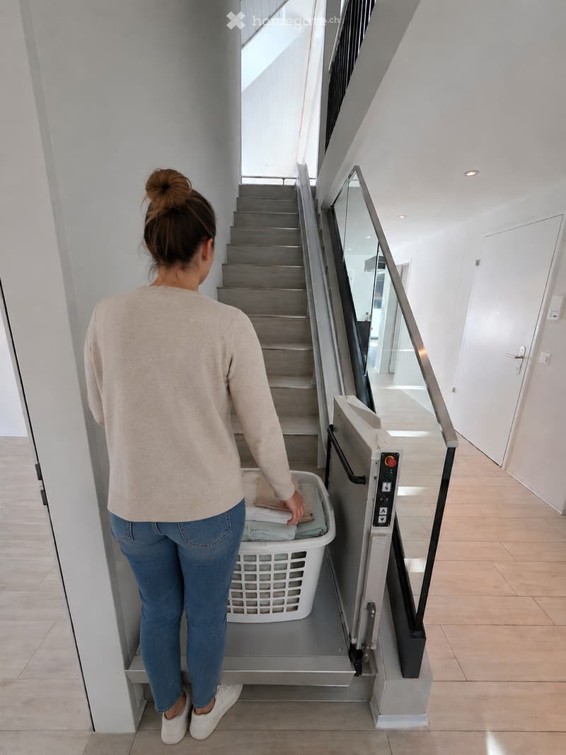 A modern hallway with a staircase, glass railing, and a laundry lift. A woman is placing laundry into the lift.