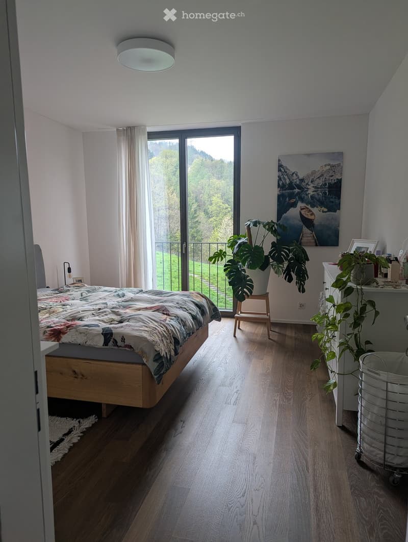 Bright bedroom with wooden flooring, large window with a view, bed with colorful bedding, nightstand, stool, plants, and artwork on the wall.
