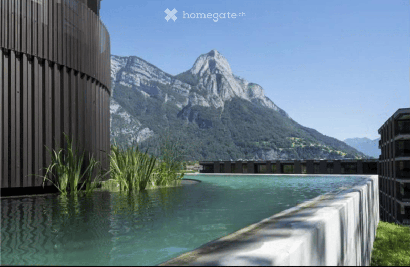 A modern outdoor pool area with a serene view of snow-capped mountains in the background, surrounded by sleek contemporary buildings.