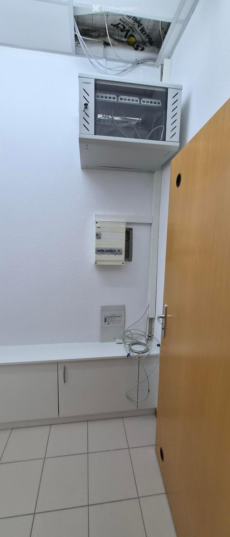 A hallway with a tiled floor, white walls, and several electrical panels and wiring visible on the wall.