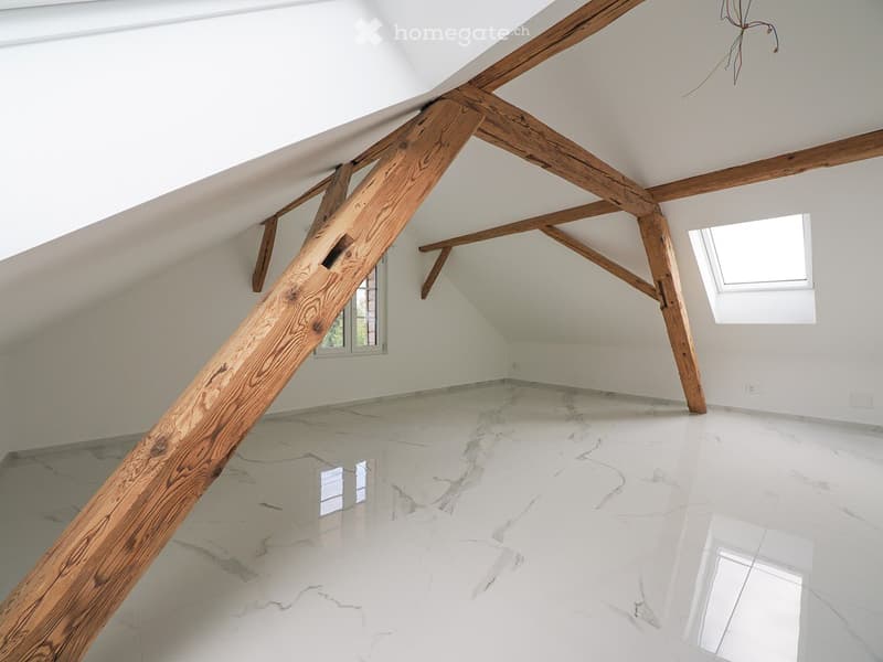 An empty attic space with exposed wooden beams, white walls, and a polished marble floor. The room features skylights and large windows, allowing natural light to fill the space.