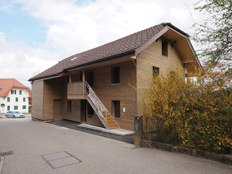 Two-story wooden house with a tiled roof, raised ground floor, and a staircase leading to a balcony.